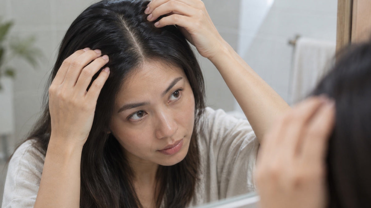 Hands gently parting dark hair to reveal a natural scalp line in a softly lit bathroom.