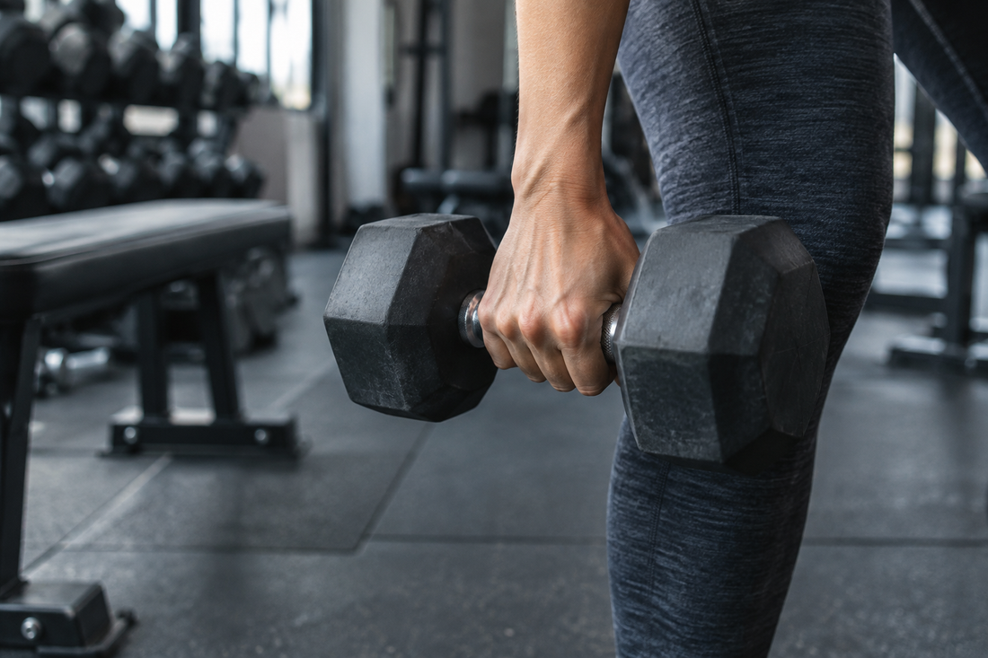A hand holding a black dumbbell beside a person's leg in a softly lit gym.